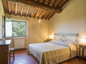 A bedroom with a wooden ceiling, a bed with a metal frame, a window with a view of trees, a radiator, and a small bedside table with a lamp. - Belvilla by OYO Piniolmo (Castelfiorentino)