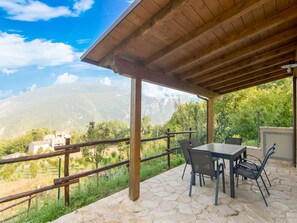 A covered outdoor seating area with a stone floor, a table with chairs, and a view of mountains and greenery.