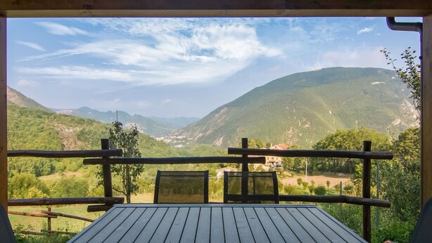 Eine Holzterrasse mit Tisch und Stühlen, mit Blick auf eine gebirgige Landschaft unter blauem Himmel.