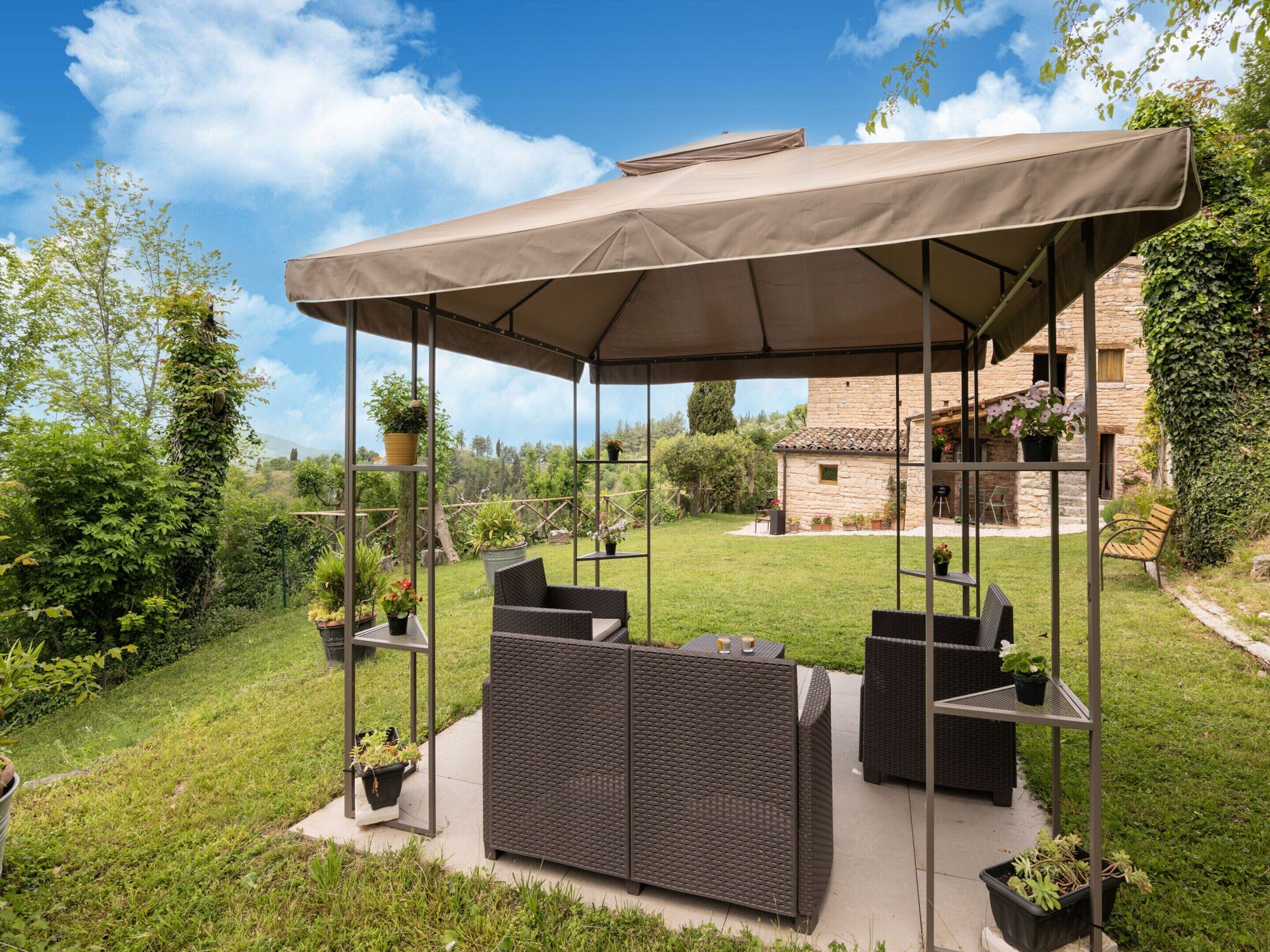 An outdoor patio with a covered seating area, surrounded by greenery and a stone building in the background.