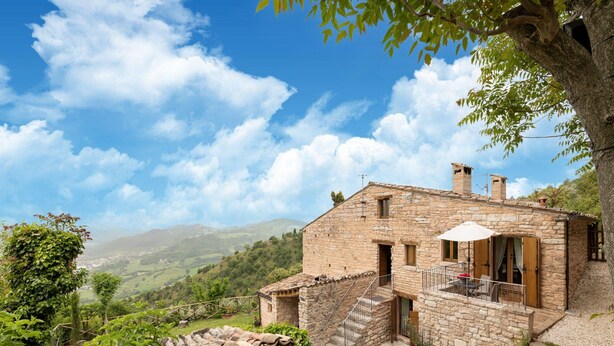 Une maison en pierre dotée d’un balcon, entourée d’une végétation luxuriante et offrant une vue panoramique sur des collines ondulantes.
