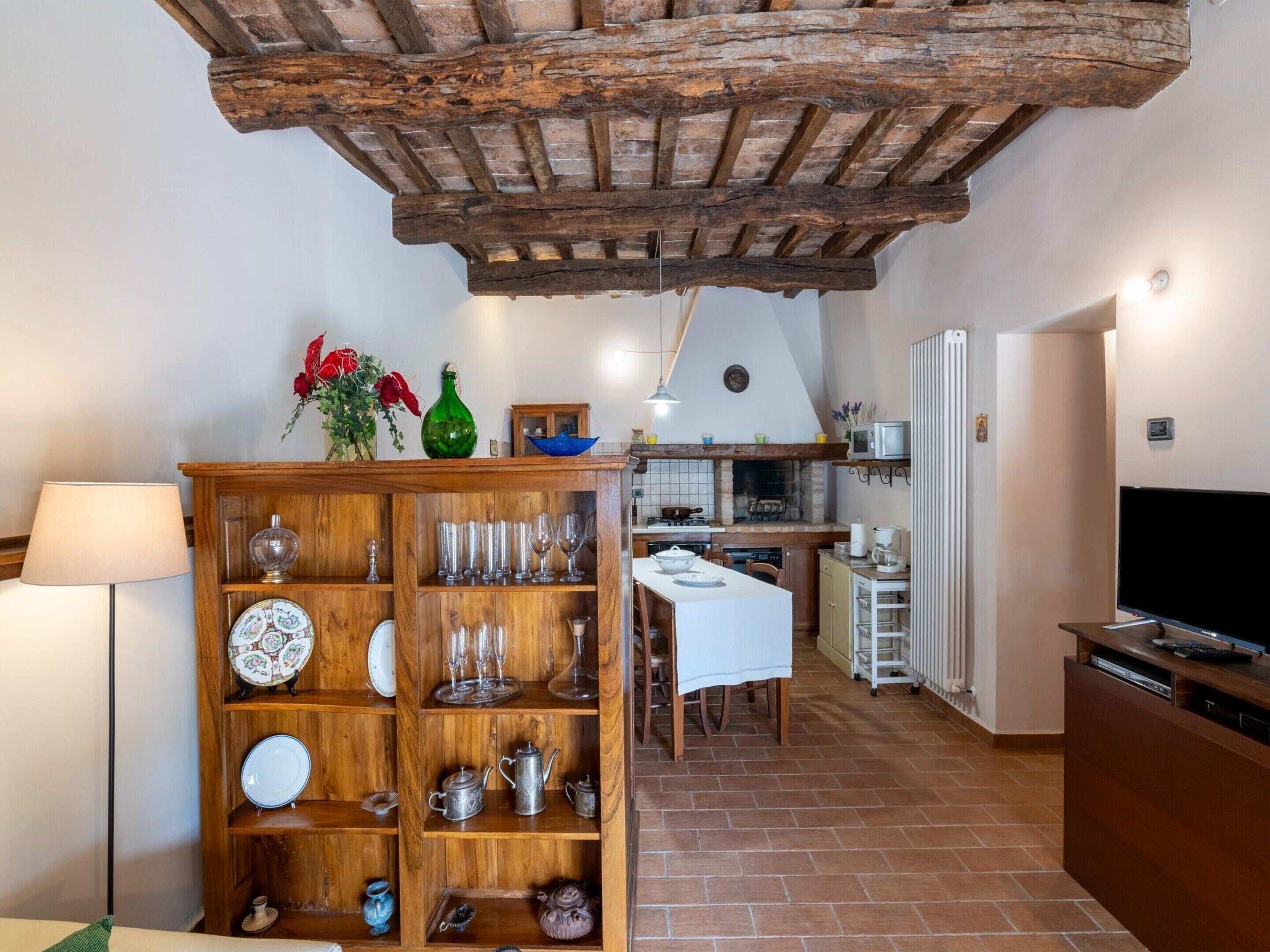 A rustic kitchen with exposed wooden beams, a dining area with a white table, and a wooden cabinet with glassware.