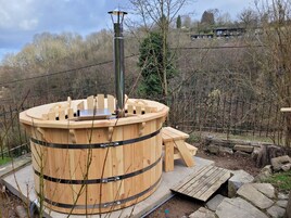 Un jacuzzi de madera con una chimenea metálica, rodeado por una terraza de piedra y un banco de madera.