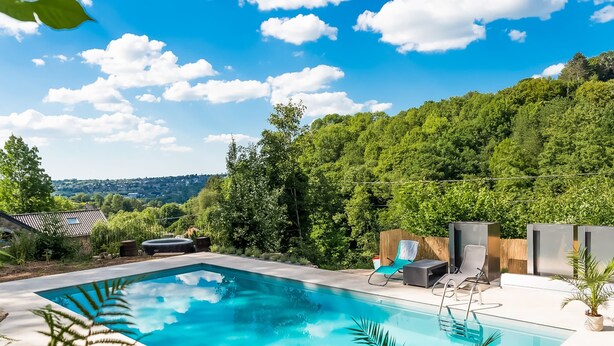 Une piscine rectangulaire à l’eau d’un bleu limpide, entourée d’une terrasse en béton. On y trouve des chaises longues et une petite table. En arrière-plan, on aperçoit une forêt verdoyante et, au loin, une ville.