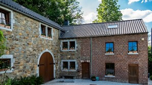 Un bâtiment en pierre et en briques, doté de nombreuses portes et fenêtres, une cour recouverte de gravier et entourée de verdure.