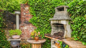 A rustic outdoor kitchen with a brick wall, a stone oven, and a wooden table surrounded by greenery. - Lovely Cottage in Vierves-sur-viroin With Garden and Terrace (Viroinval)