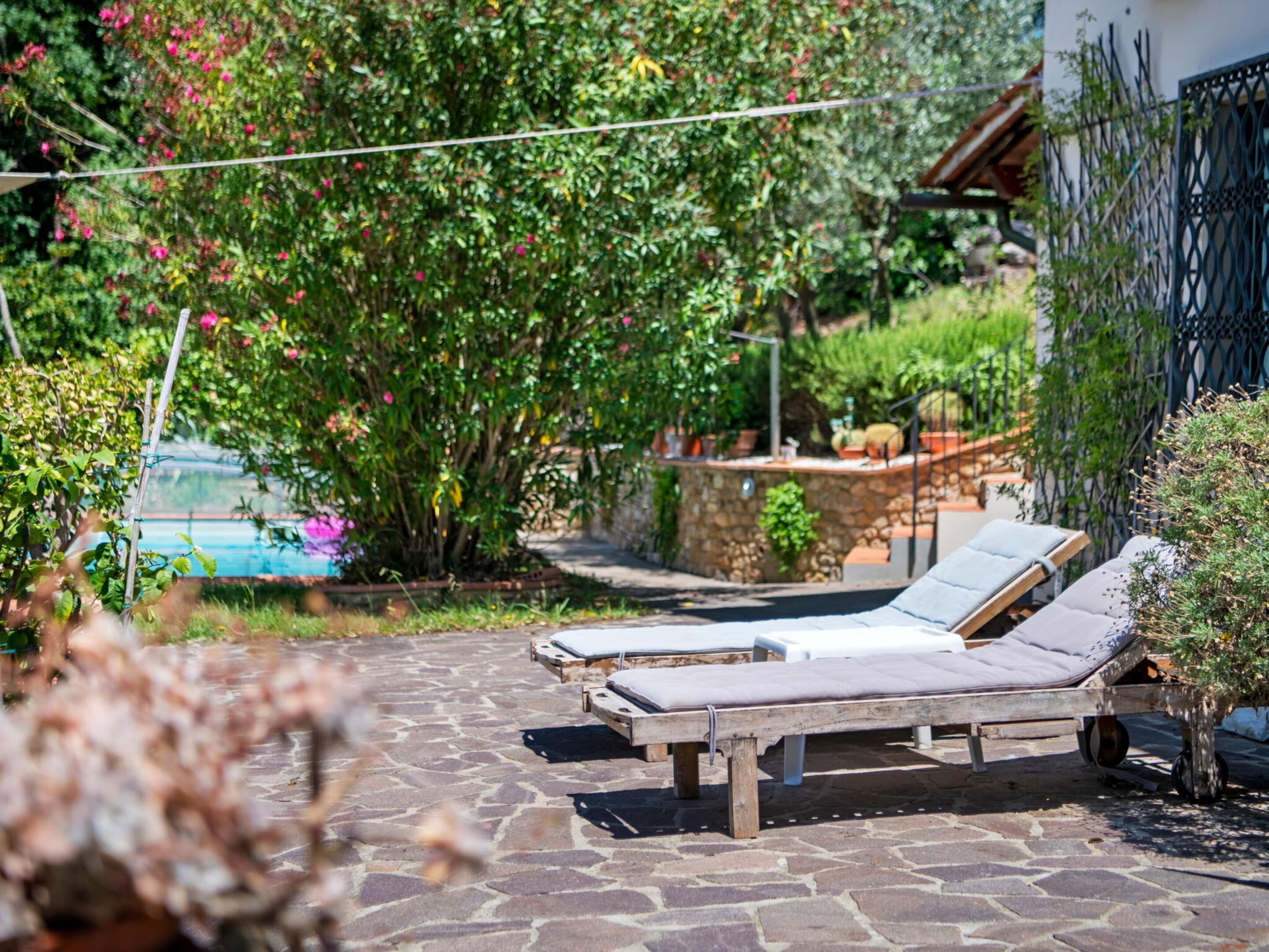 A stone patio with two sun loungers, a stone wall, and lush greenery.