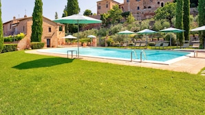 A swimming pool with lounge chairs and umbrellas, surrounded by well-maintained grass and trees, with buildings in the background. - Belvilla by OYO Il Melograno (Montalcino)
