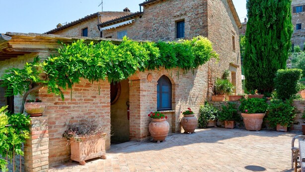 A courtyard with a brick building, potted plants, and a pergola covered with greenery. - Belvilla by OYO Il Melograno (Montalcino)