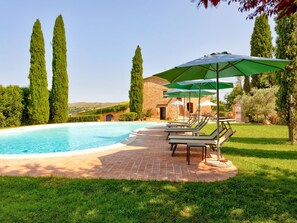 Zona de piscina con sillas de descanso y sombrillas, rodeada de follaje y un edificio de piedra.