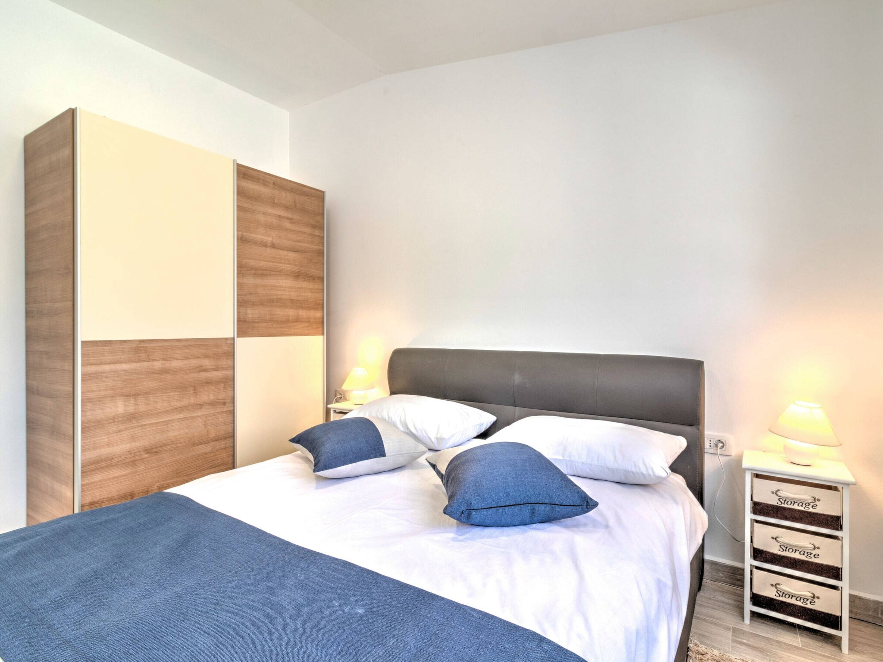 A neatly made bed with a grey headboard, white bedding, and blue pillows. There are two bedside tables with lamps, and a tall storage unit with drawers labeled "Storage" on the side.