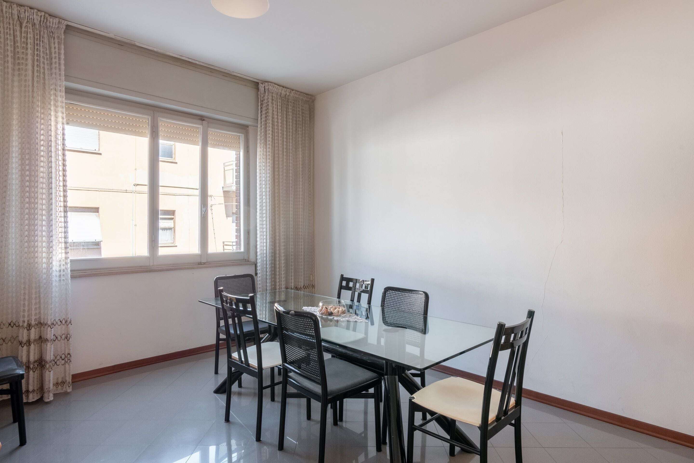 A dining area with a glass table and black chairs, a window with curtains, and a plain white wall.