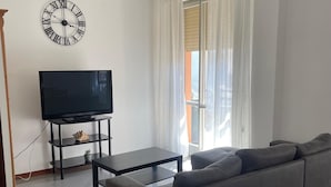 A living room with a sofa, a TV on a stand, a clock, and a window with curtains. - Holiday Home in Fano Near the Beach (Fano)
