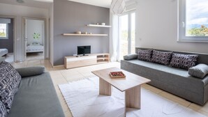 A modern living room with a grey sofa, a coffee table, a TV on a wooden stand, and a view of the outdoors through a large window. - Lovely Apartment in Novalja near Sea Beach & Town Center (Novalja)