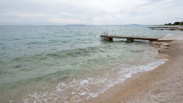 A pebbled beach with a wooden pier extending into calm waters, surrounded by distant hills. - Lovely Apartment in Novalja near Sea Beach & Town Center (Novalja)