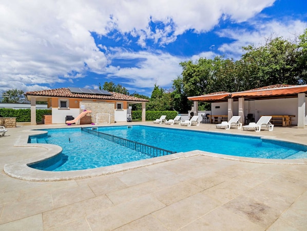 A swimming pool with a sun terrace, lounge chairs, and a covered area with a table and chairs.