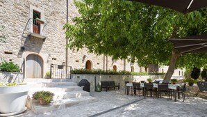 Une cour avec des marches en pierre menant à un espace aménagé avec des tables et des chaises, le tout entouré d’un bâtiment en pierre doté de fenêtres en arc de cercle.