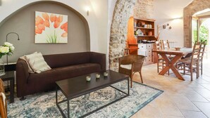 Un salon chaleureux avec un canapé marron, une table basse et un tableau représentant des fleurs oranges. On y trouve un mur en pierre et une arche en bois qui mène à un coin cuisine avec des meubles en bois.