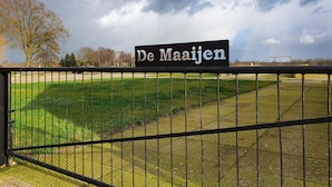 Een bord met de tekst 'De Maijen' voor een groen veld en een bewolkte lucht.