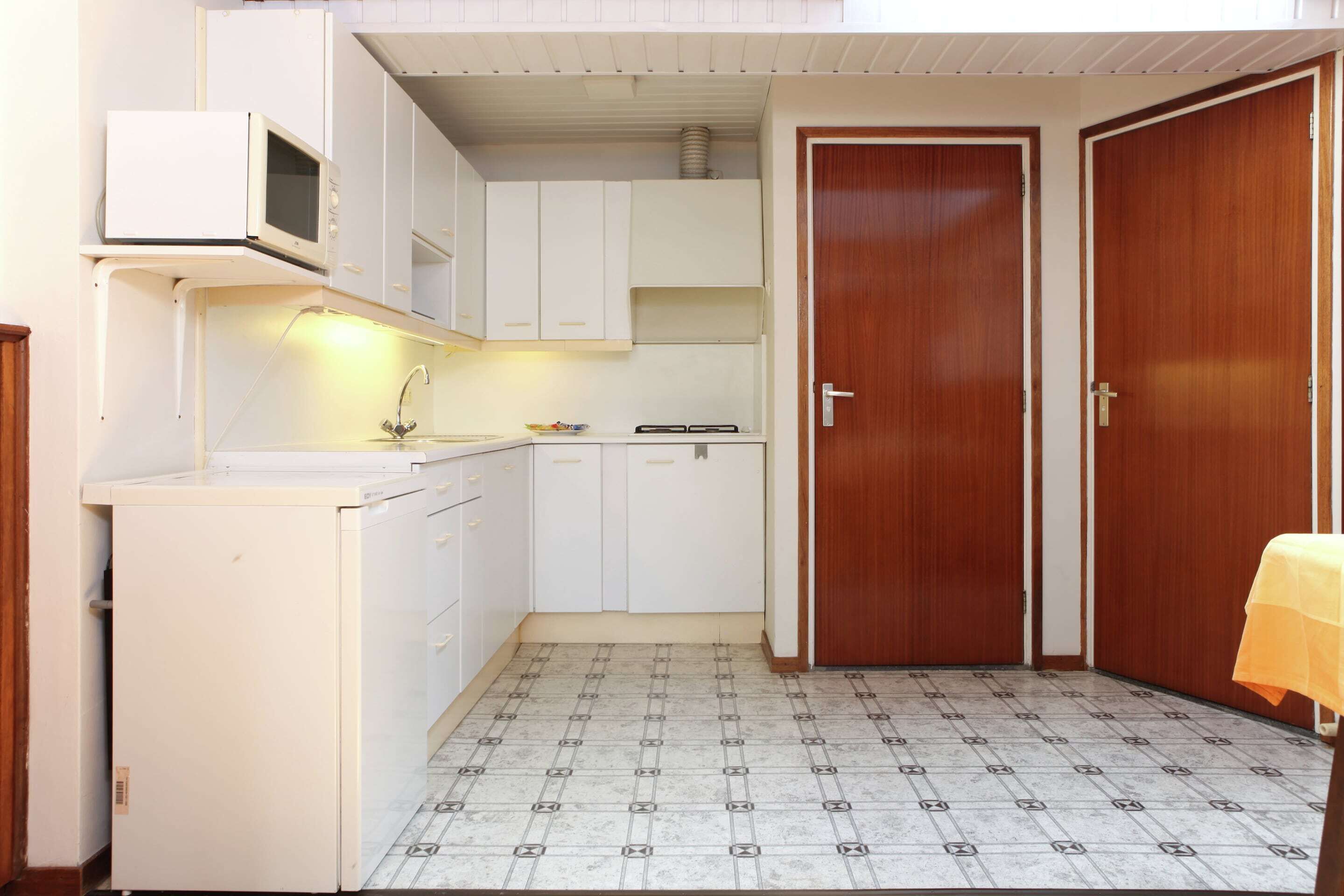 Une cuisine compacte avec des meubles blancs, un four à micro-ondes, un évier et une cuisinière. On y trouve une porte en bois et une table recouverte d’une nappe jaune.