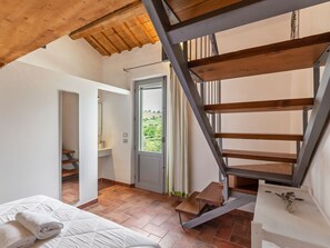 Camera da letto con soffitto in legno, un letto con biancheria da letto bianca, bagno con lavandino e specchio, e una scala che conduce a una zona esterna.