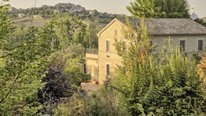 A rustic stone building surrounded by lush greenery, with a distant view of a hilltop town.