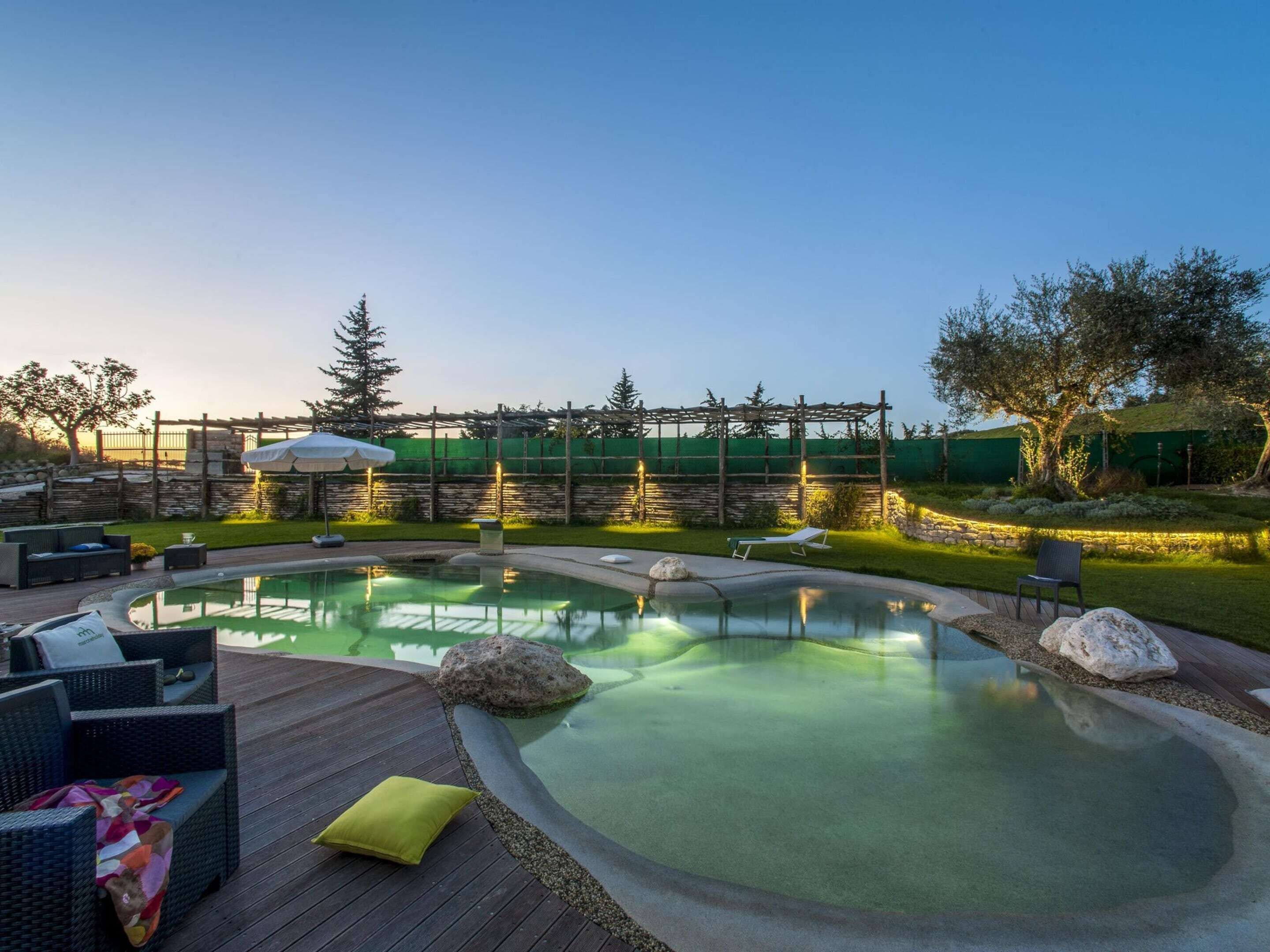 Una piscina con bordo in legno, poltrone relax e un pergolato con luci decorative.