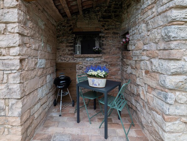 A stone-walled outdoor seating area with a table and chairs, a grill, and a flower arrangement.