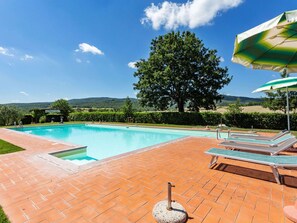 A swimming pool with a view of hills, surrounded by a patio with lounge chairs and an umbrella.