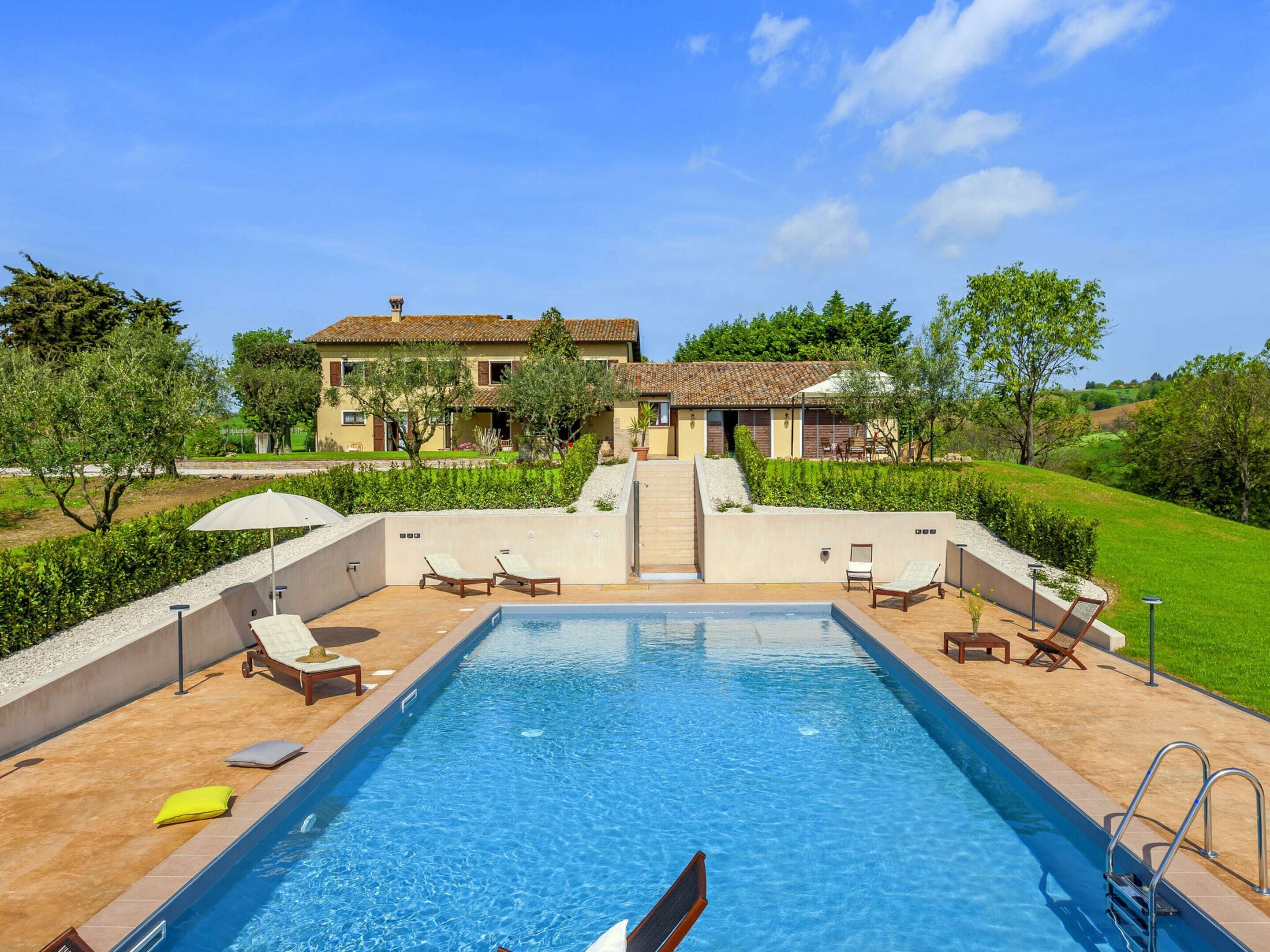 A rectangular swimming pool with a clear blue water surface, surrounded by a tiled deck. There are lounge chairs and umbrellas along the poolside.