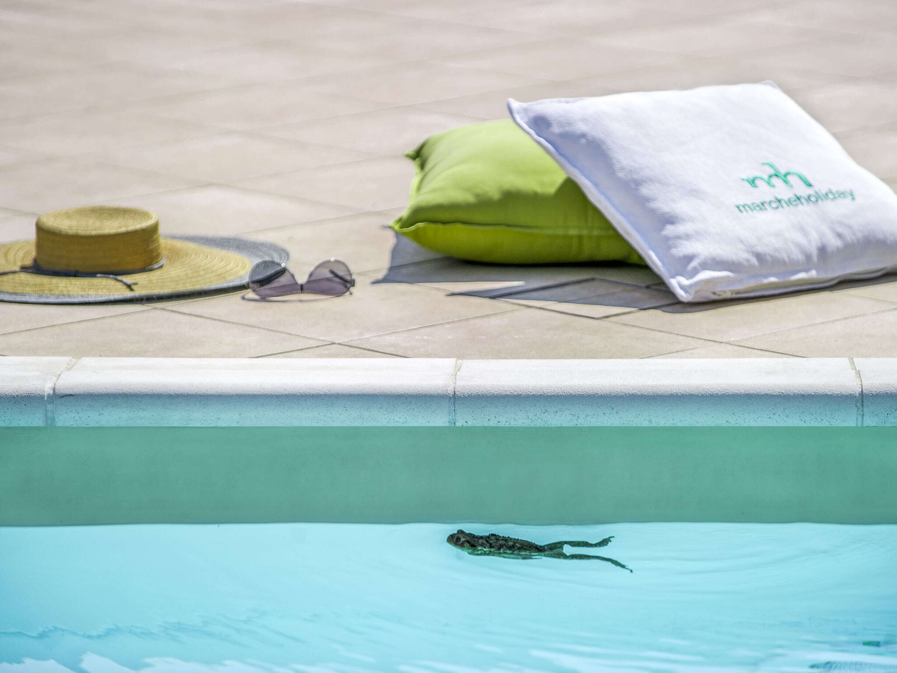 A poolside area with a green pillow, a white towel, a straw hat, and sunglasses on the ground.