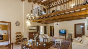Amplia sala de estar con una mesa de comedor de madera, sillas, un sofá y un televisor. Una escalera conecta con un nivel superior.