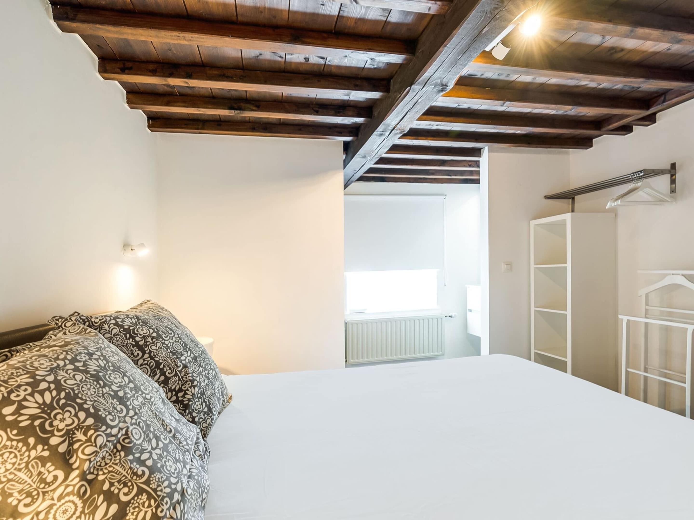 A bedroom with a wooden ceiling, a bed with patterned pillows, a white radiator, and a white wardrobe with a hanging rod.