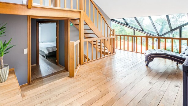 A spacious room with a wooden staircase leading to an upper level, a large window with a view of greenery, and a modern black leather sofa.