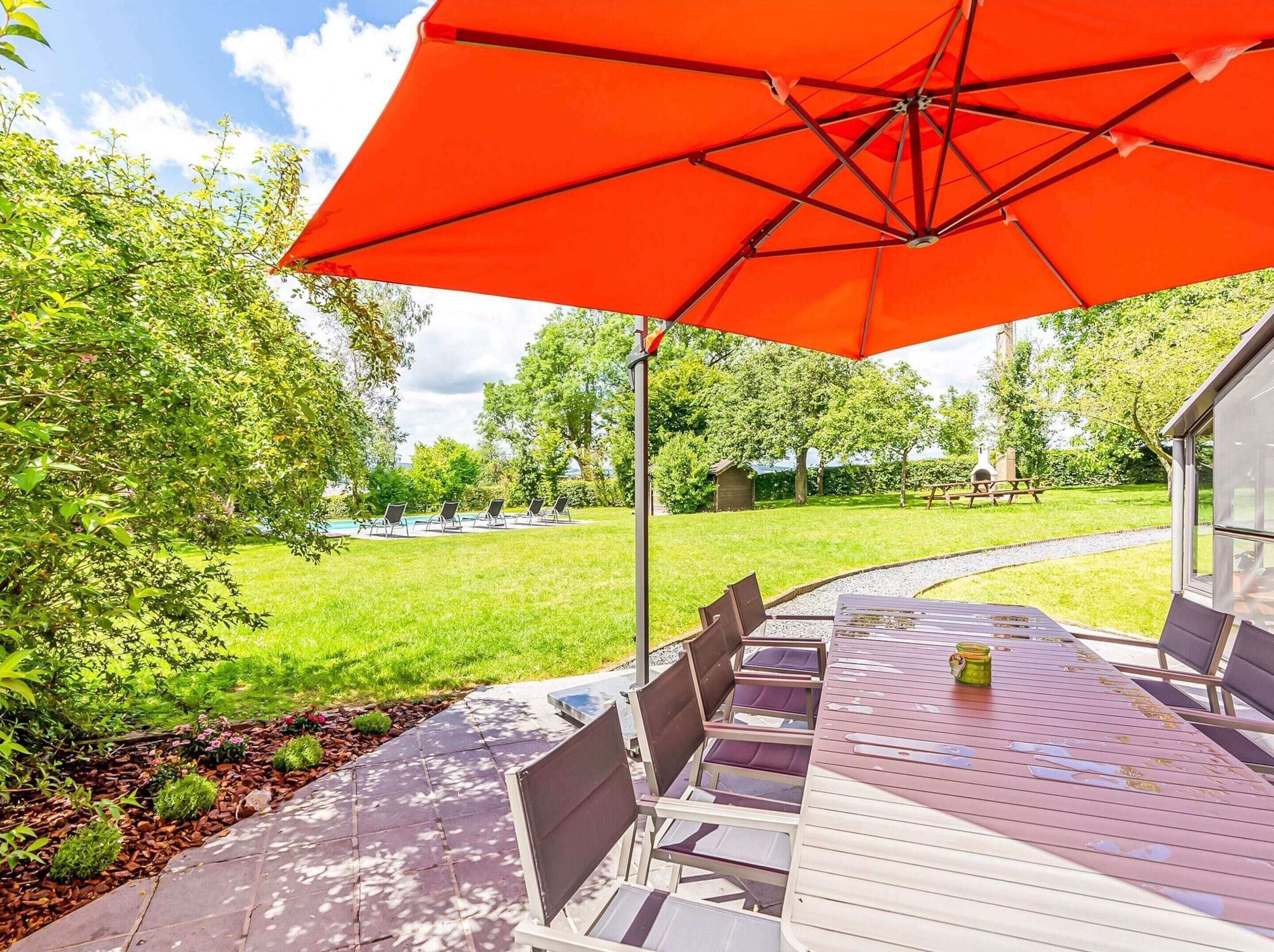 An outdoor patio with a table and chairs under a large red umbrella, surrounded by greenery and a view of a swimming pool.