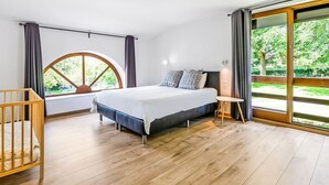 A modern bedroom with a large bed, a wooden crib, a small table, and a large window with a view of greenery.