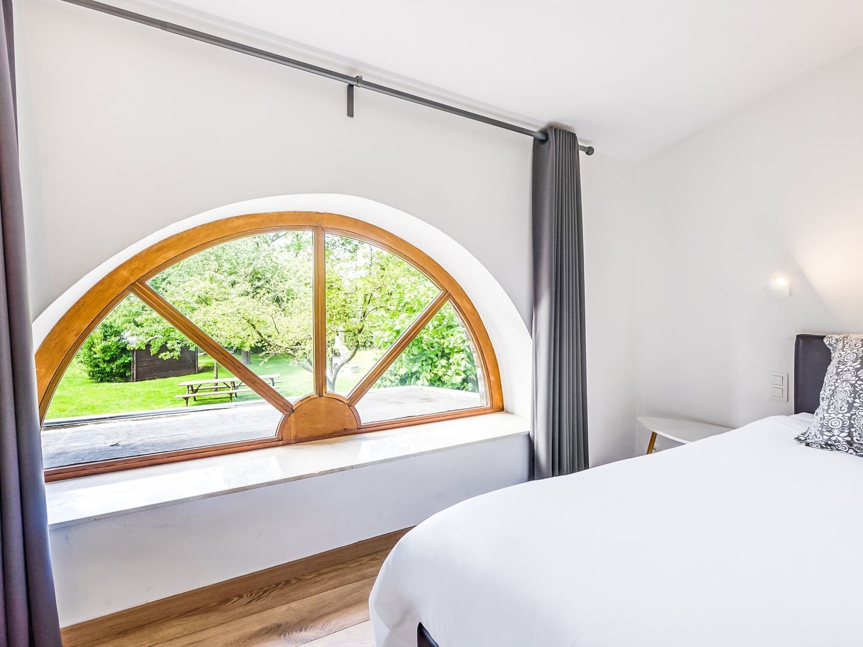 A bedroom with a large arched window, a bed, and a view of a garden.