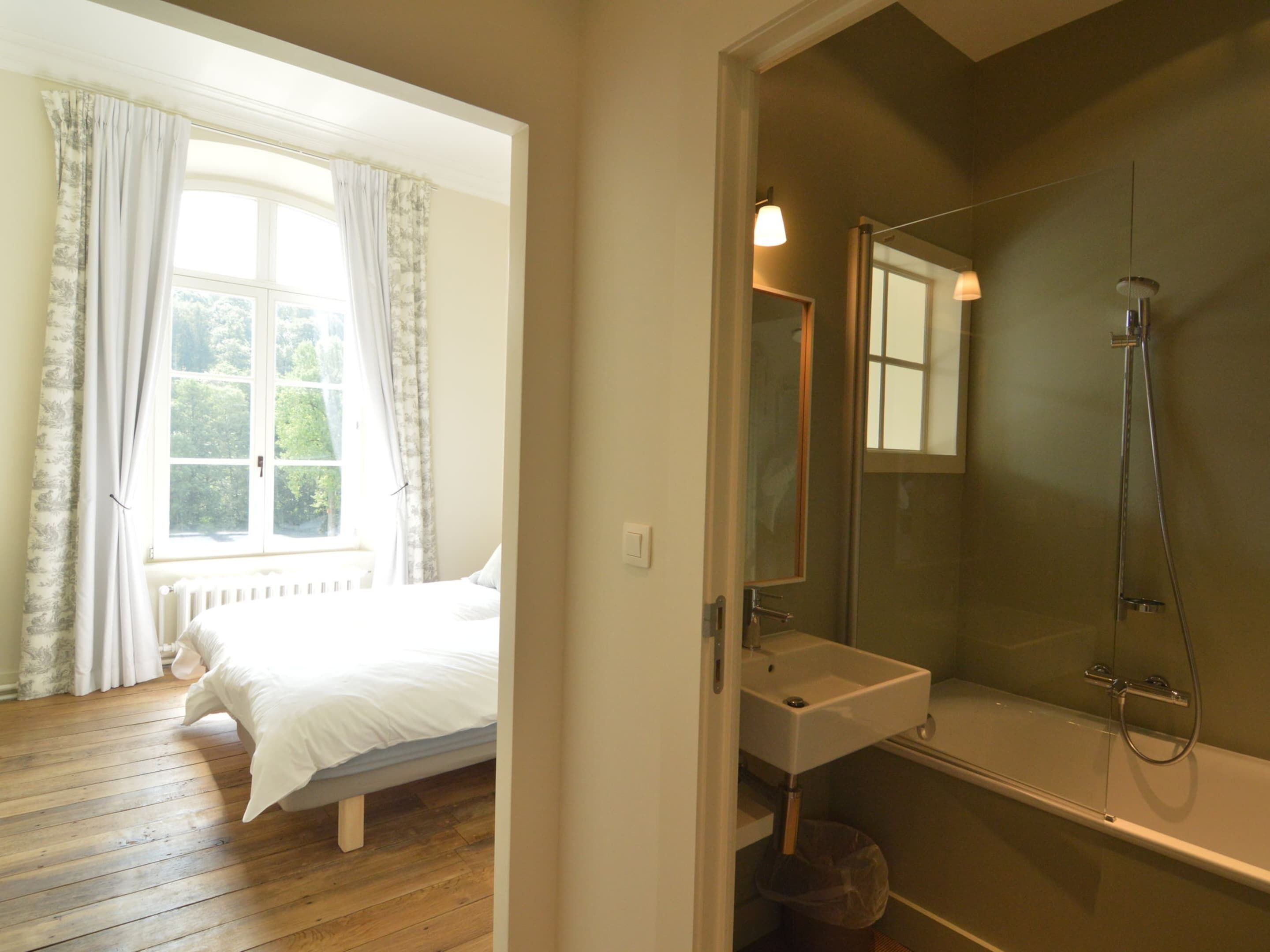 A bedroom with a wooden floor, a bed, a window with curtains, and a bathroom with a sink and shower.