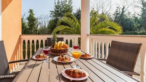 A wooden table set with croissants, a fruit basket, and drinks on a balcony with white railings and palm trees in the background. - Alluring Apartment in Banjole near Beach & Island (Medulin)