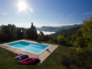 A rectangular swimming pool with a tiled edge, surrounded by grass and trees, with a view of mountains and a lake in the background. - Eco-retreat With Pool (Acqualagna)