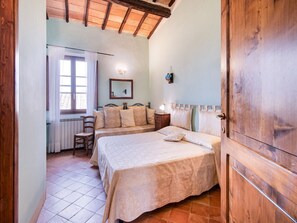 A bedroom with two beds, a wooden ceiling, a window with curtains, a bedside table with a lamp, and a chair.