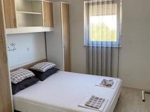 A neatly made bed with a pillow and a folded towel, a window with curtains, and a wall with a heart and word design.