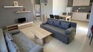 A modern living room with a grey sofa, a wooden coffee table, and a TV. There is a kitchen area with cabinets and a dining table. - Apartment in Novalja Near Pag Beaches (Novalja)