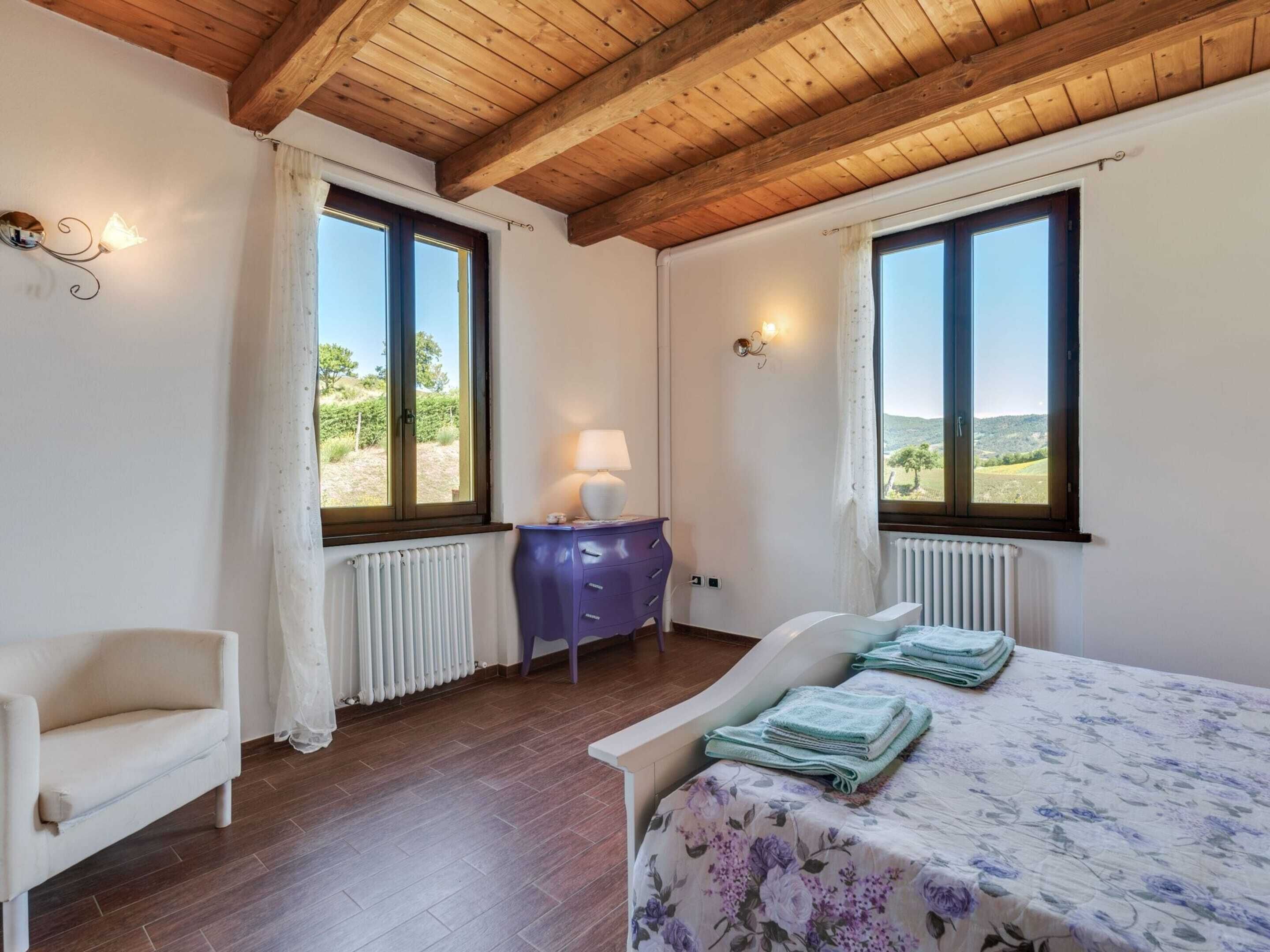 A bedroom with a wooden ceiling, two windows with curtains, a bed with a floral bedspread, a chair, a small table with a lamp, and a radiator.