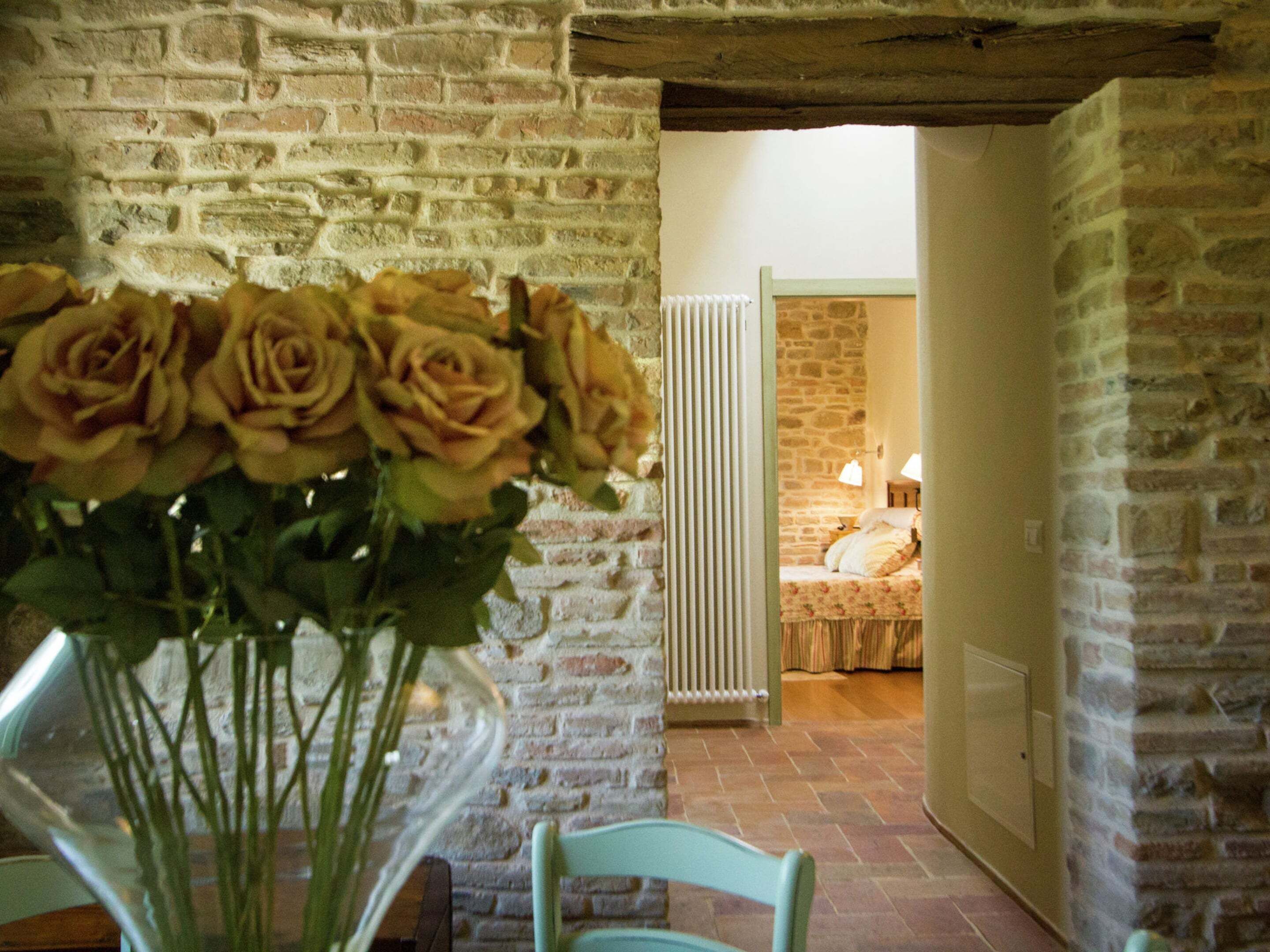 Une pièce avec un mur en briques, un lit recouvert d’une courtepointe à motifs floraux et un vase de roses jaunes posé sur une table.