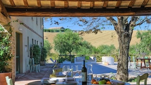 An outdoor dining area with a table set for two, surrounded by chairs, a potted plant, and a view of a hillside.