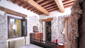 Une chambre avec un mur en pierre, des poutres apparentes en bois au plafond, un lit avec du linge de lit blanc, une commode et un miroir.