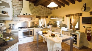 A rustic kitchen with a stone wall, wooden beams, a stove, and a dining table set with food. - Belvilla by OYO La Rocca (Apecchio)