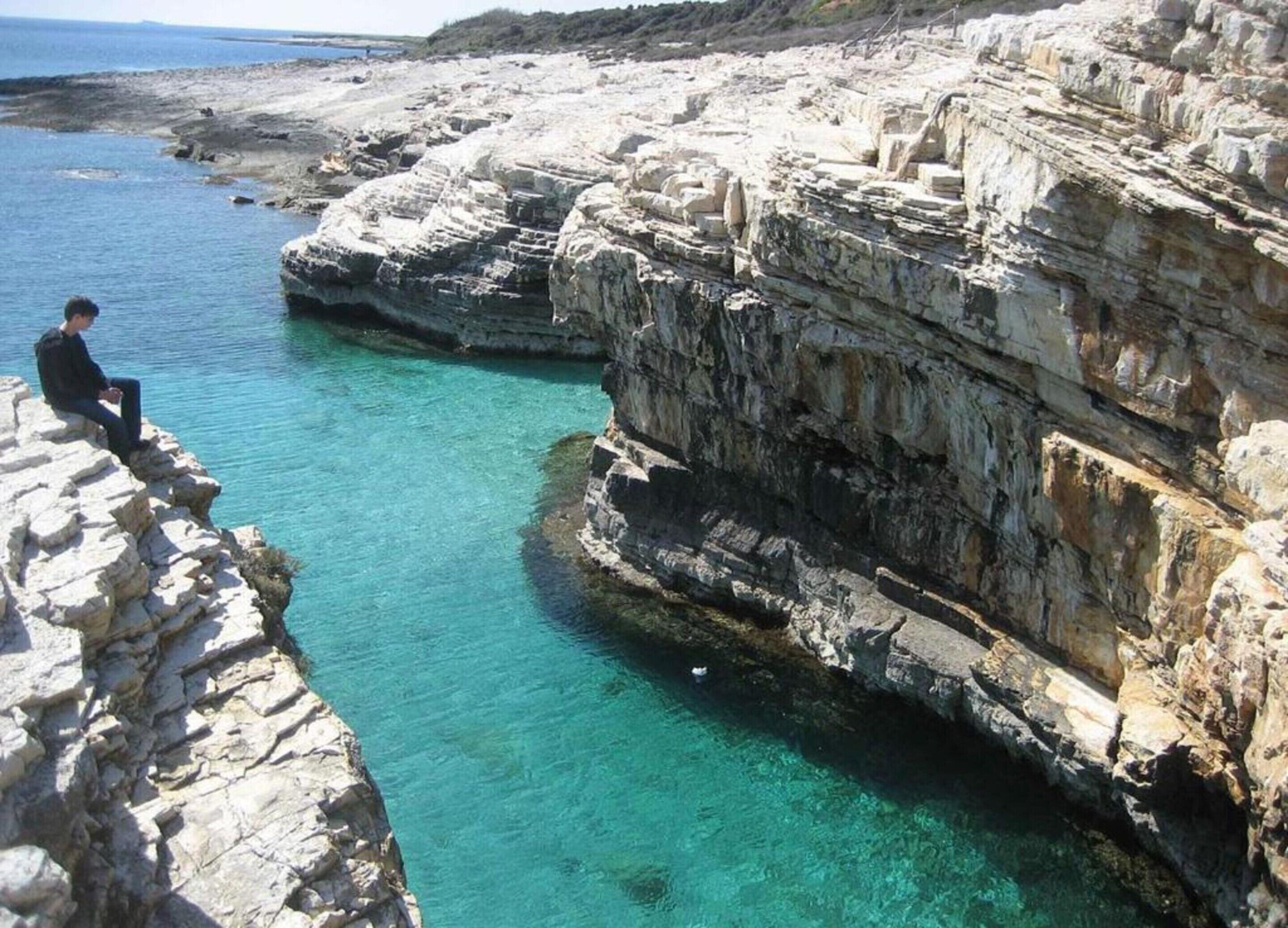 A rocky coastal area with clear blue water and a person sitting on a rock.