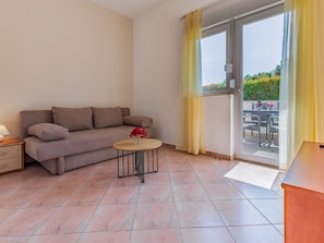 Uma sala de estar com sofá, uma mesinha redonda com um vaso de flores e vista para um pátio externo através de uma janela grande.